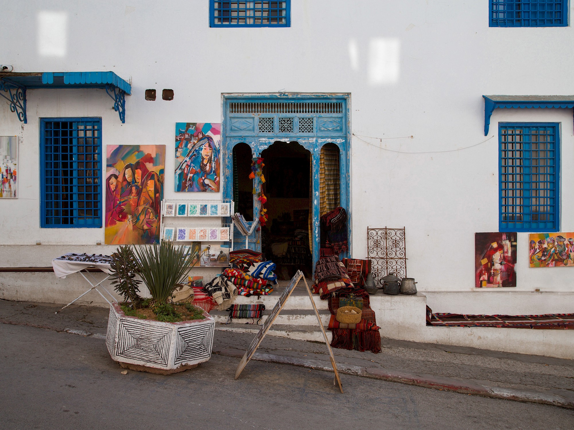 A whitewashed shopfront with light-blue trim on a cobbled lane, framed paintings and patterned rugs propped up outside.