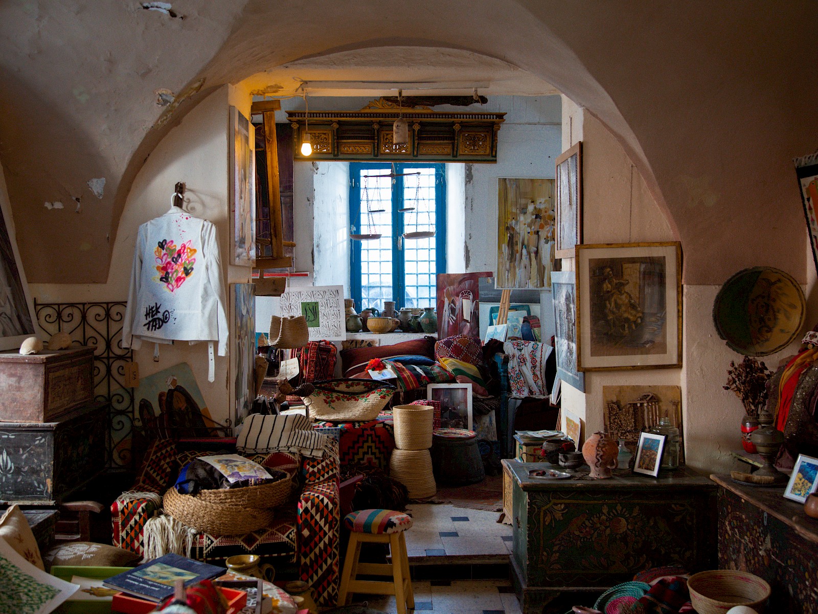 The interior of a crowded art shop with white vaulted ceilings, walls of framed paintings and stacked rugs, and a big blue window casting sunlight at the back.