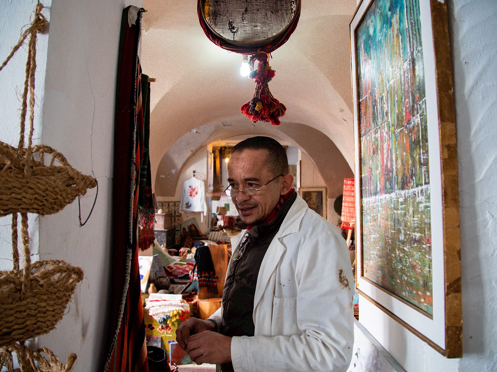 A middle-aged man in a white coat stands looking down in the cluttered shop, hanging crafts on one side and framed paintings on the other.