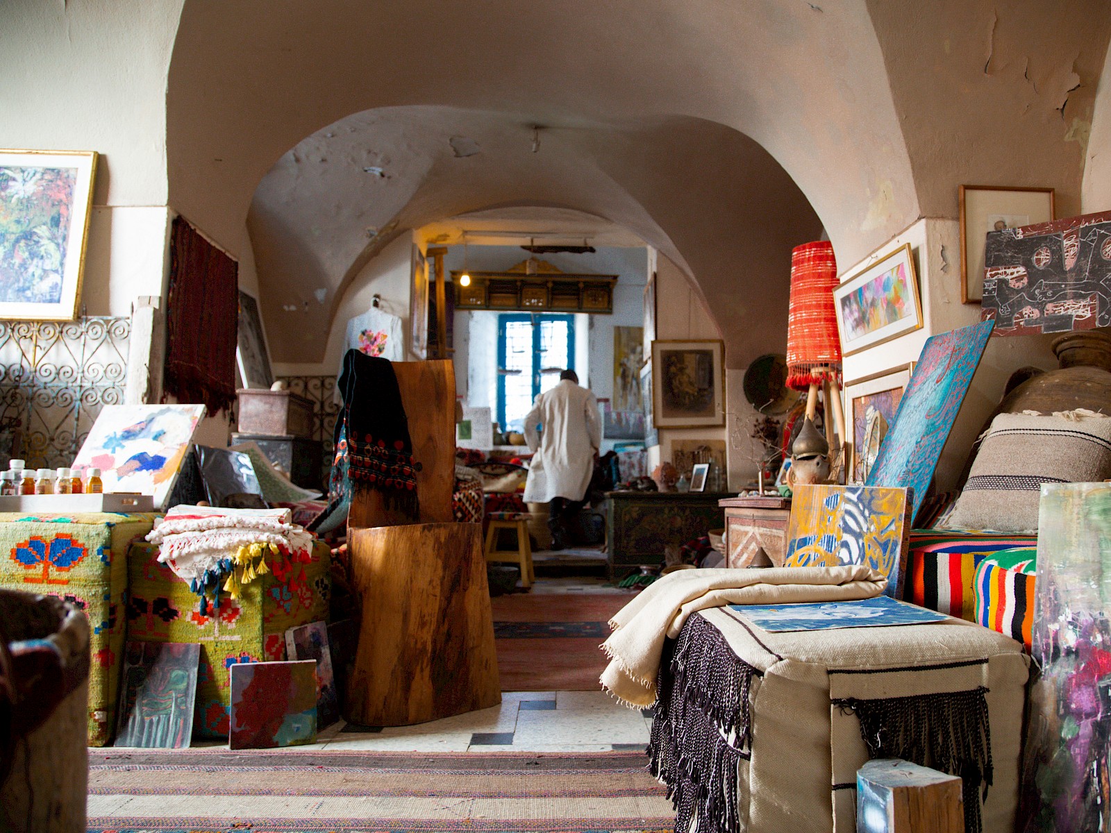 A narrow path through piles of textiles and canvases, leading toward a bright window at the back of the shop.