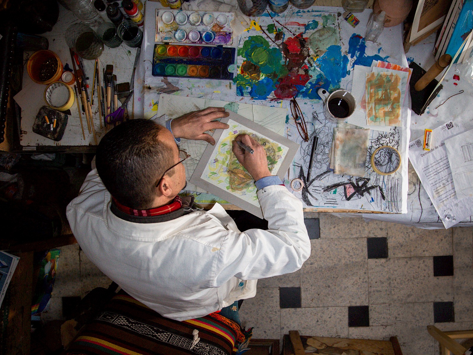 Overhead view of the man in the white coat leaning over a cluttered desk, focused on a colourful drawing.