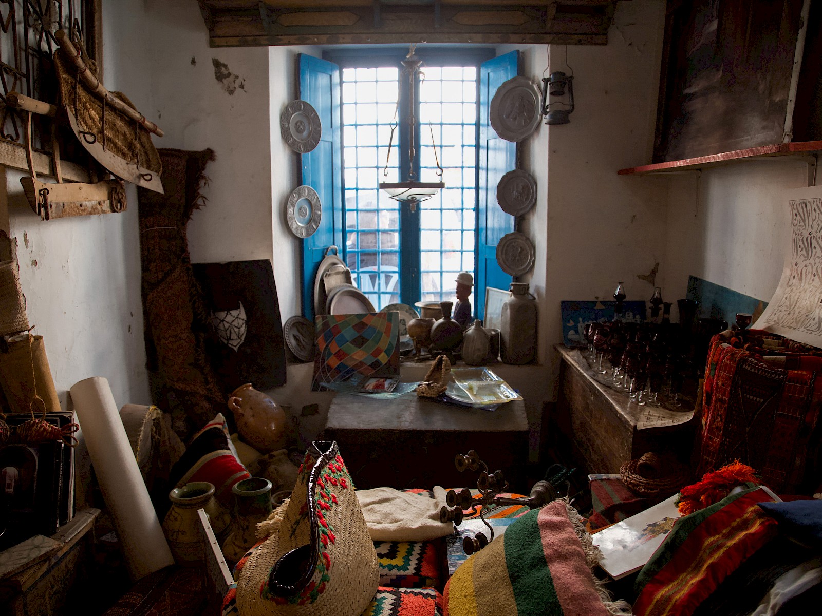 A shop window with blue wrought-iron grilles; bright daylight behind it turns the pottery and crafts in the foreground into dark silhouettes.