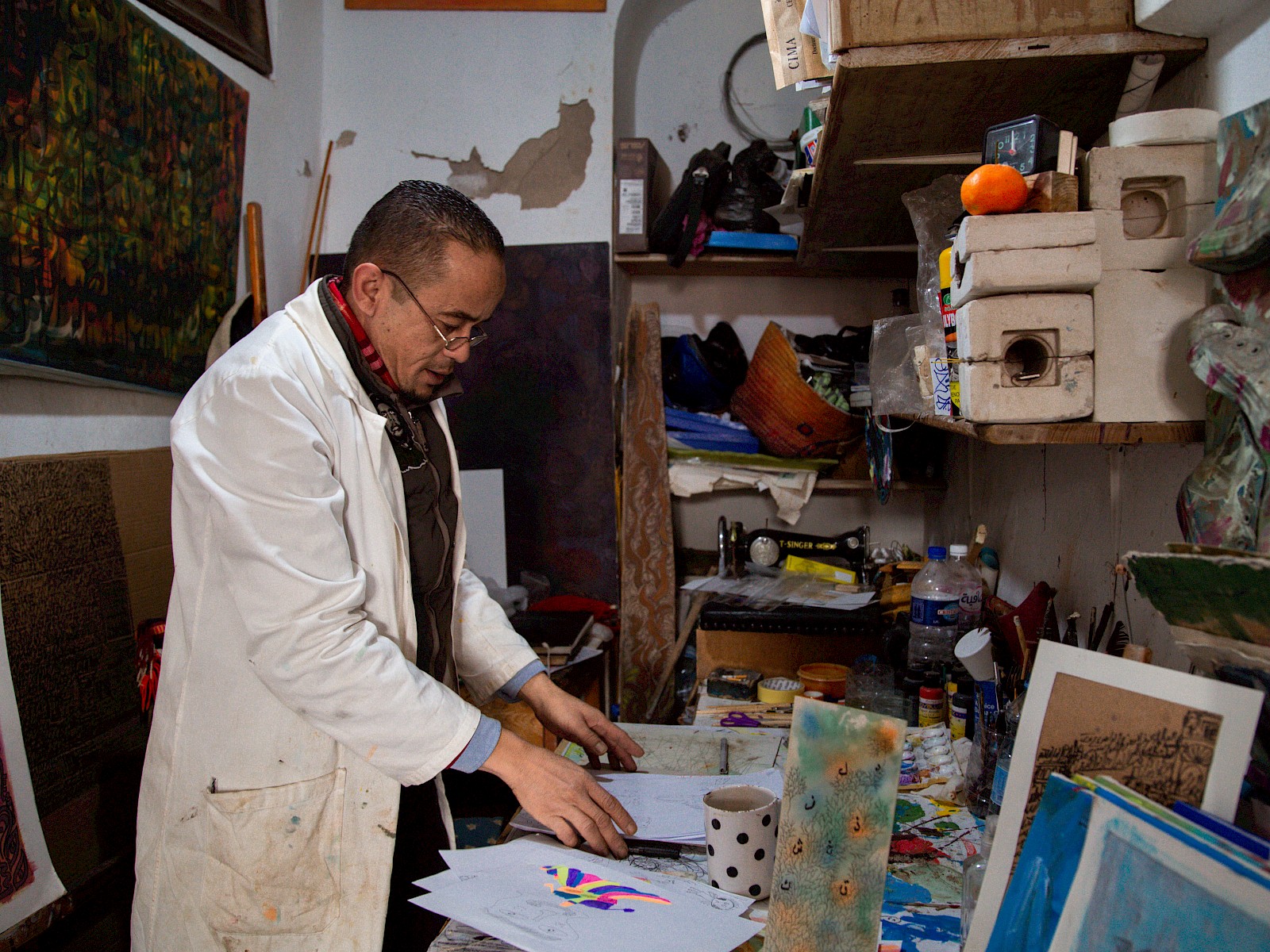 The man stands at a counter sorting sheets of artwork, light from the right lighting his face and white coat against the dark room.