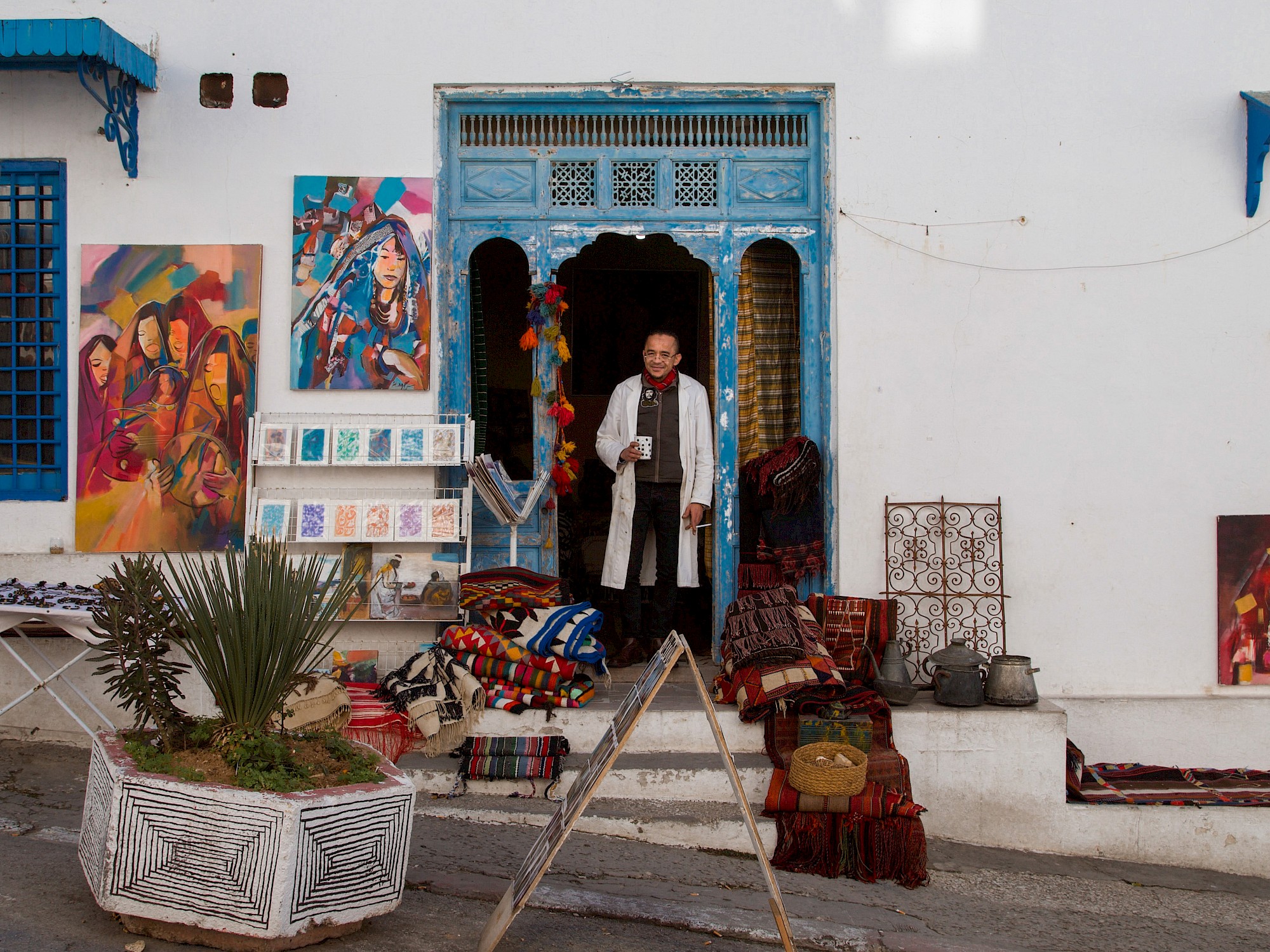 The man stands in the open blue double doors of the shop, facing the street with a slight smile.
