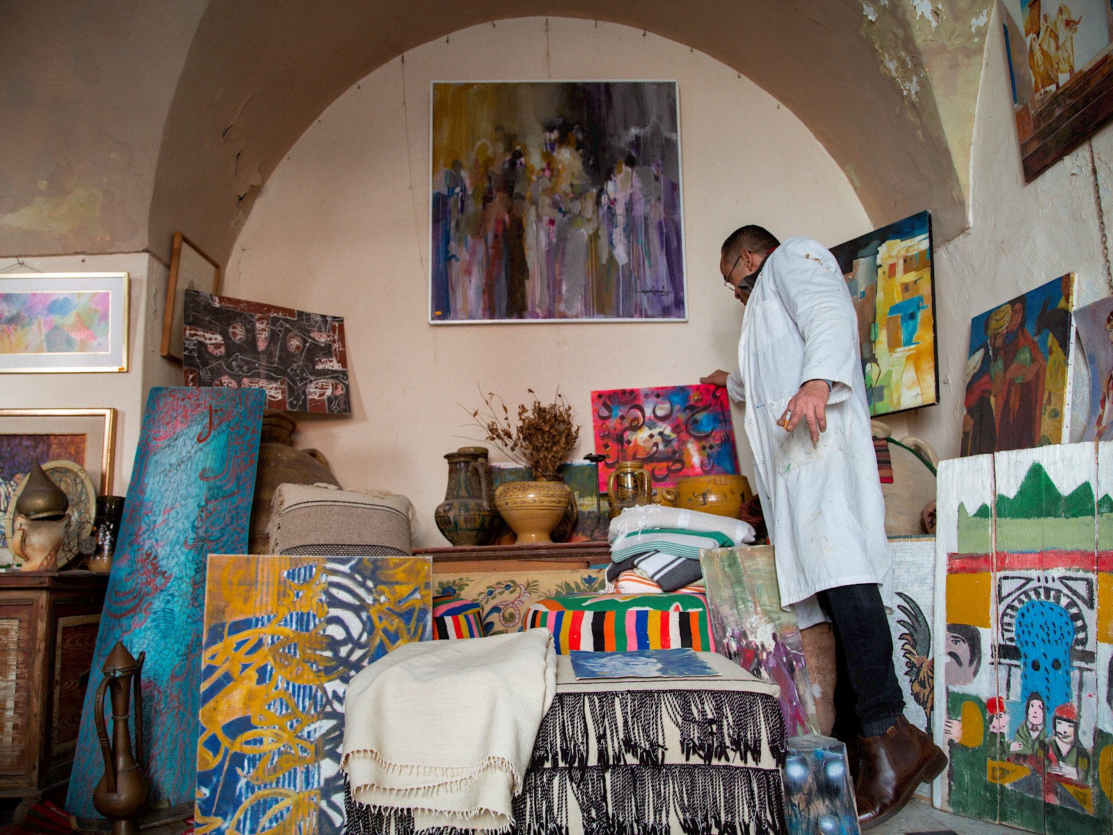 The man stands beside an easel holding a brightly patterned painting, with more canvases stacked against the wall behind him.